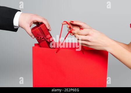 vista ritagliata dell'uomo che mette la scatola del regalo in borsa di carta nelle mani di donna con labbra rosse isolate su grigio Foto Stock