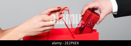 vista ritagliata dell'uomo che mette la scatola del regalo in sacchetto di carta nelle mani della donna con le labbra rosse isolate sul grigio, striscione Foto Stock