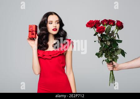 uomo che dà rose alla donna di brunette in vestito rosso che tiene confezione regalo confezionata isolata in grigio Foto Stock