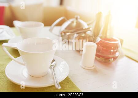 Colazione o tè nel ristorante. Tavolo in caffetteria. Tazza con piattino, cucchiaio, teiera di tè, sale, pepe, zucchero e vaso con rosa sul tavolo. Foto Stock