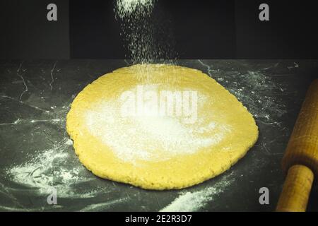 Pasta sfoglia sul tavolo. L'impasto grezzo viene arrotolato su una superficie di lavoro infarinata con un perno di laminazione in legno Foto Stock