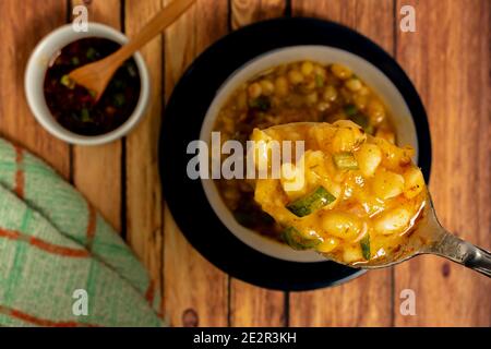 Primo piano di un cucchiaio con locro. Sfondo sfocato di una ciotola bianca con tipica cucina argentina locale e la salsa piccante che accompagna questo stufato Foto Stock