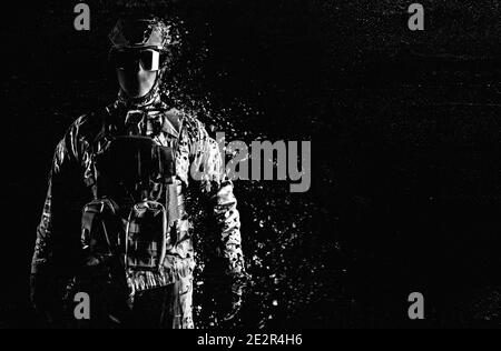 Foto in bianco e nero di un soldato completamente attrezzato in piedi in abiti tattici e sciogliersi su sfondo nero grugnoso. Foto Stock