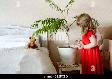 Carino ragazza del bambino dei capelli rossi nella camera da letto che innaffia una pianta della casa in un letto vicino al letto. Cura esotica della pianta, giardinaggio domestico Foto Stock