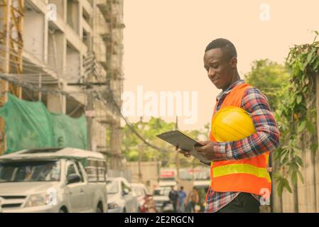 Giovani felici Africano nero uomo lavoratore edile sorridere mentre la lettura sulla clipboard e azienda elmetto in cantiere Foto Stock