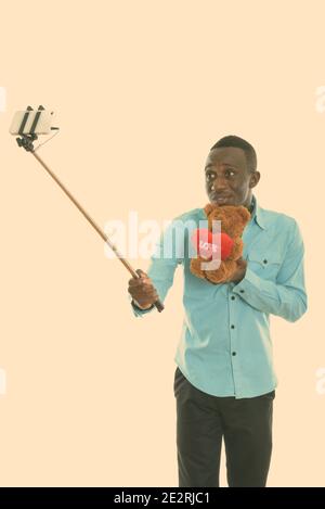 Giovane nero africano holding orsacchiotto di peluche con cuore e amore segno tenendo selfie foto con il telefono cellulare su selfie stick Foto Stock