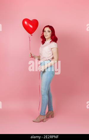 giovane donna snella e felice a tutta lunghezza con i capelli rossi in jeans tiene un grande palloncino rosso volante a forma di cuore, isolato su uno sfondo rosa Foto Stock