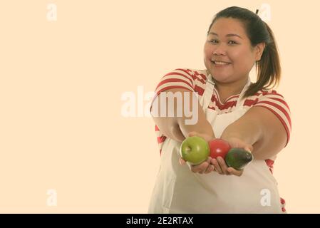 Studio shot di giovani felici fat donna asiatica sorridere mentre dando frutti freschi e indossando il grembiule Foto Stock