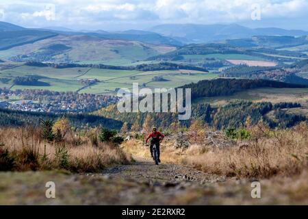 Un uomo guida una mountain bike elettrica in salita lungo una strada ghiaiosa nelle colline sopra la città di Peebles nei confini scozzesi. EBike Scotland. Foto Stock
