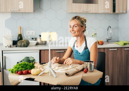 Una donna carina cucina in cucina e guarda una ricetta video al telefono o comunica tramite chat video. Foto Stock