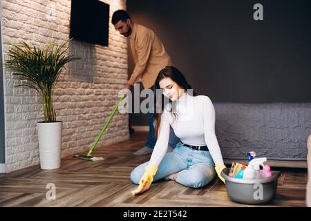 La famiglia giovane pulisce il loro appartamento a casa. L'uomo attraente sta mescolando il pavimento con la donna carina. Concetto di pulizia e pulizia della casa. Foto Stock