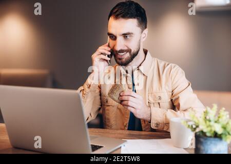 Lavoratore maschile remoto di successo. Giovane freelance felice di una telefonata. Cucina moderna sullo sfondo. Programmatore freelance maschio che lavora da casa Foto Stock