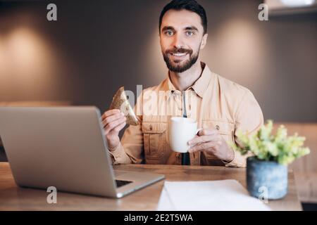 Uomo freelancer brunette che fa colazione e guarda la macchina fotografica. Più caffeina. Uomo in abiti casual utilizzando il computer portatile, la programmazione. Concetto di lavoro remoto Foto Stock