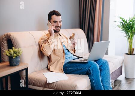 Lavoro a distanza durante il concetto di blocco. Happy maschile freelance si siede sul divano nel suo salotto e parla con un cliente utilizzando il telefono cellulare guardando il compito Foto Stock