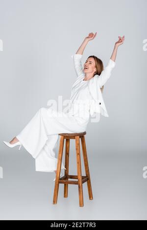tutta la lunghezza di donna eccitata in vestito bianco seduto sopra sgabello su grigio Foto Stock