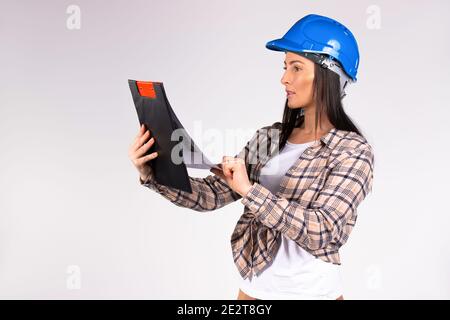 Giovane ingegnere donna in un casco blu legge il piano di lavoro. Sfondo grigio e lavori di architetto femminile in corso. Foto Stock