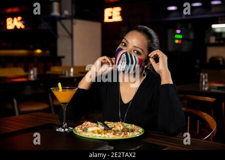 Ritratto di una donna seduta in un ristorante mentre indossa Covid 19 facemask protezione, concetto di allontanamento sociale, mangiare cibo. Foto Stock