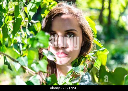 Una bella giovane donna che si nasconde dietro foglie verdi e verdi illuminate dal sole Foto Stock