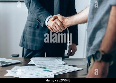 Vista ritagliata di handshaking investitore con uomo d'affari in primo piano offuscato vicino a computer portatile e carte in primo piano sfocato Foto Stock