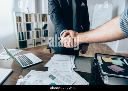 Vista ritagliata di uomo d'affari scuotendo mano di investitore vicino a documenti con grafici e dispositivi sul tavolo Foto Stock