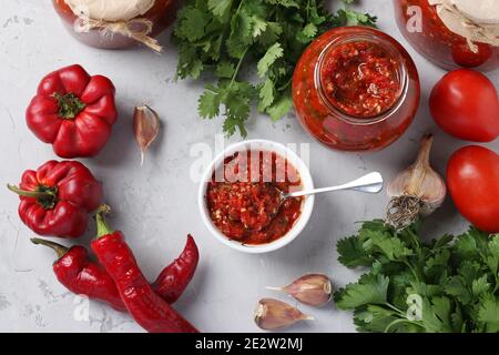 Adjika armena di pepe rosso, pomodori, aglio, coriandolo e prezzemolo in una ciotola e vasi su sfondo grigio. Vista dall'alto Foto Stock