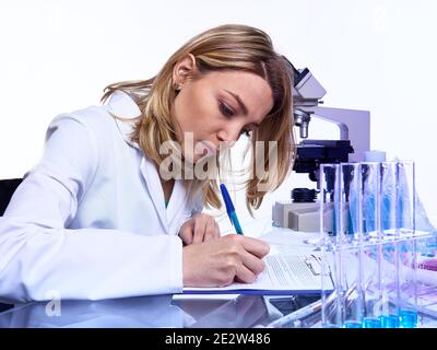 Una giovane scienziata femminile o un tecnico scrive report o un giornale di laboratorio in un laboratorio moderno con microscopio, provette in vetro con campioni e timer, immagine tonata al neon Foto Stock