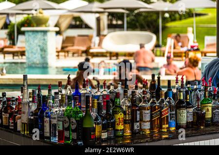 Numerose bottiglie di liquori allineano il bancone di un bar in piscina presso un hotel turistico vicino a St. George's, Grenada, comprese le marche chiaramente leggibili Jameson, Dewar's e Jack Daniel's. Lo sfondo sfocato mostra gli ospiti che si rinfrescano in piscina. Foto Stock