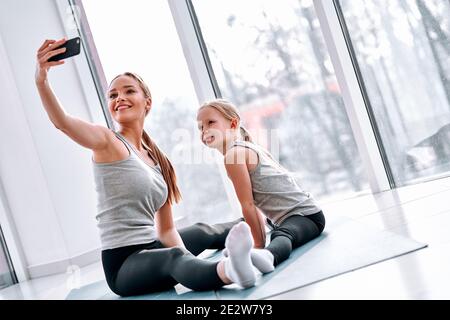 Giovane mamma sport blogger fare selfie con la sua bambina carina mentre facendo il riscaldamento, esercizi di stretching in classe yoga. Concetto di blog di sport per la famiglia Foto Stock