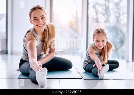 Mamma e figlia stanno facendo yoga. Famiglia in palestra. Bambina con madre in una maglietta grigia e leggings neri. Ragazze che si siedono su un tappetino e si allungano Foto Stock