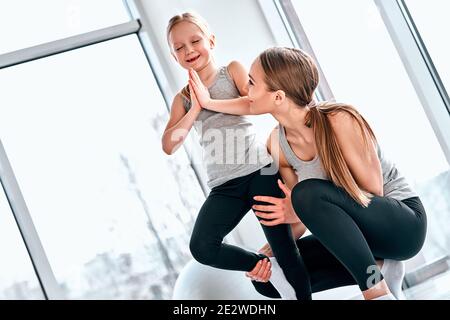 Ritratto di madre che insegna bambina piccola con gli occhi chiusi facendo esercizio di yoga, aiutando la figlia a tenere la gamba in posa albero, asana nel fitness cl Foto Stock