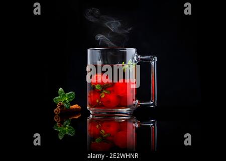 Tè alla ciliegia calda con cannella e menta in una tazza di vetro su tavolo a specchio e sfondo nero Foto Stock