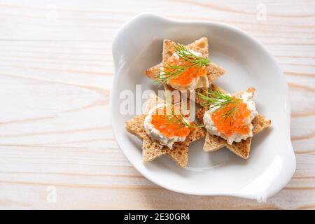 Canapes con caviale rosso su pane tostato a forma di stella con crema e guarnizione di aneto su un piatto bianco, tavolo di legno chiaro, spazio di copia, vista ad alto angolo f Foto Stock