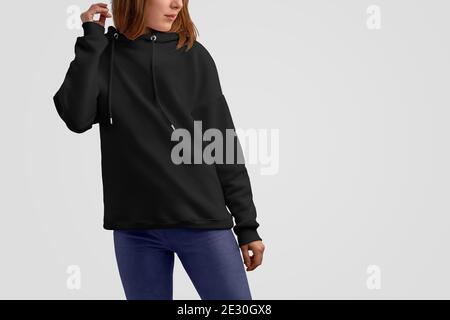 Serie di abiti mockup. Donna caucasica gracile in una felpa nera con cappuccio e pantaloni blu isolati sullo sfondo vuoto dello studio. Posa frontale. Modello CAN u Foto Stock