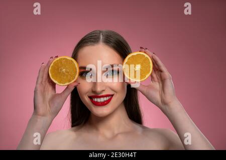 Ritratto di una giovane donna positiva con arance affettate in mani che si presentano affascinanti. Affascinante gioiosa signora divertente con lunghi capelli isolati su rosa Foto Stock