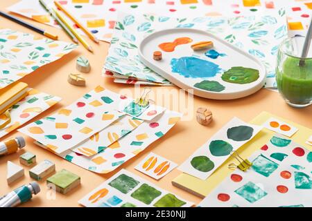 Fogli di carta con campioni di vari ornamenti geometrici disposti vicino tavolozza con colori e strumenti colorati su sfondo arancione Foto Stock
