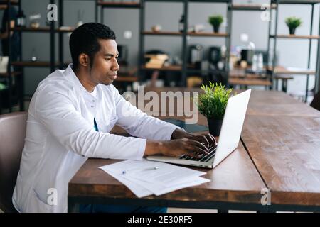 Focalizzato giovane medico africano maschio in bianco cappotto di lavoro digitando sul computer portatile seduto alla scrivania. Foto Stock