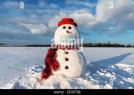 Simpatico pupazzo di neve in elegante cappello rosso con maschera medica su campo innevato. Concetto di allontanamento sociale e di viaggio annullato Foto Stock