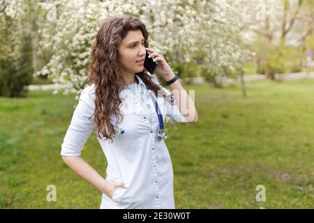 dottore di sesso femminile che parla su cellulare . sfondo medico spazio copia . all'aperto di un ospedale in giardino fiorito . Giovane allegra donna . Foto Stock