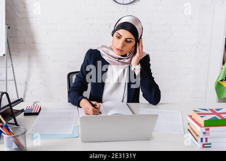 interprete arabo in cuffia scrittura in notebook vicino al portatile, traduttore digitale e dizionari di lingue straniere Foto Stock