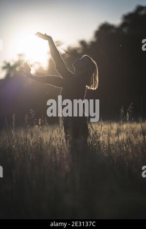 Una giovane donna danzava nell'erba alta, retroilluminata dal sole che tramonta durante l'ora d'oro Foto Stock