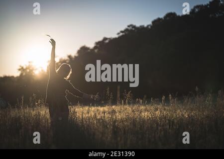 Una giovane donna danzava nell'erba alta, retroilluminata dal sole che tramonta durante l'ora d'oro Foto Stock