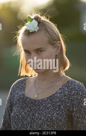 Una donna dai capelli biondi indossa un fiore bianco nei capelli, retroilluminato dal sole che tramonta durante l'ora d'oro Foto Stock