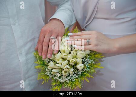 Gli sposi hanno messo le mani su un bouquet di rose bianche di fidanzamento. La sposa ha messo la mano sulla mano del marito con gli anelli di nozze Foto Stock