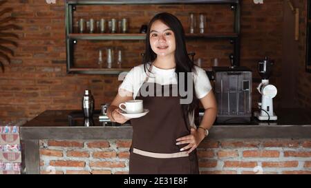 Asian Women Barista sorridente e guardando la macchina fotografica nel banco della caffetteria. Barista femmina che tiene una tazza di caffè al caffè Foto Stock