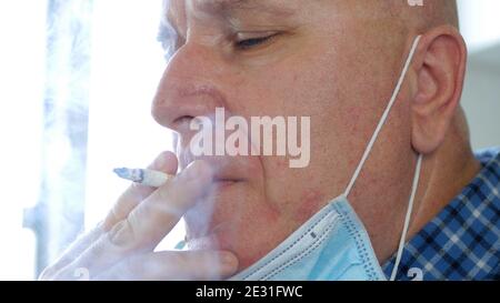 Immagine con un uomo che fuma una sigaretta utilizzando su La faccia una maschera protettiva contro l'infezione da virus Foto Stock
