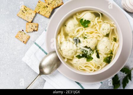 Zuppa con polpette di pollo e pasta d'uovo, parmigiano, prezzemolo in un recipiente in ceramica su uno sfondo grigio. Brodo italiano tradizionale. Vista dall'alto. Foto Stock