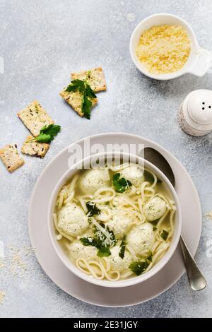 Zuppa con polpette di pollo e pasta d'uovo, parmigiano, prezzemolo in un recipiente in ceramica su uno sfondo grigio. Brodo italiano tradizionale. Vista dall'alto. Foto Stock