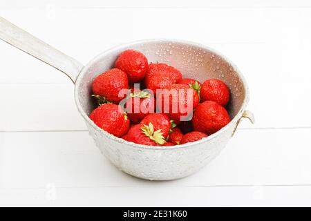 Fragole mature rosse appena lavate in un vecchio colander di metallo con gocce d'acqua su tavolo di legno bianco Foto Stock