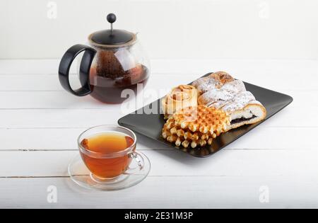 Tazza di tè, piattino con pasticceria e teiera rotonda trasparente su tavolo di legno bianco. Foto Stock