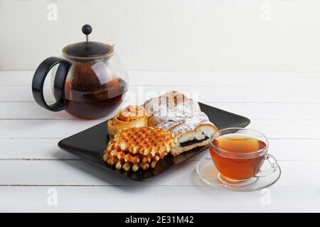 Tazza di tè, piattino con pasticceria e teiera rotonda trasparente su tavolo di legno bianco. Foto Stock